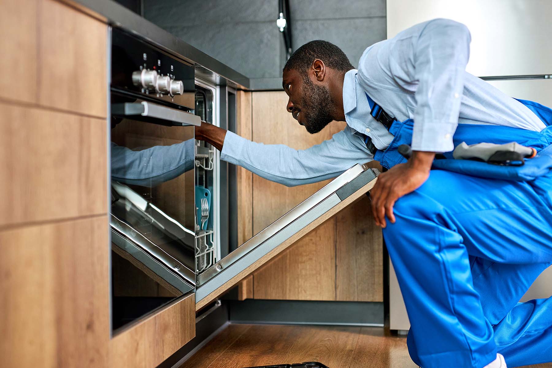 A technician in blue overalls inspects a dishwasher.